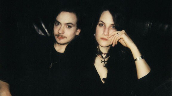 Ein Mann und eine Frau in schwarzer Kleidung sitzen auf einem schwarzen Ledersofa. 