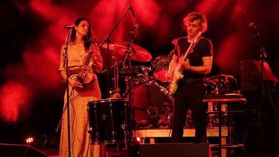 Ein Bühne, auf der linken Seite steht eine Frau in langem Kleid mit einer Violine, rechts ein Mann mit einer Gitarre. 