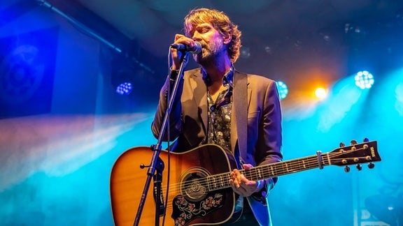 Niels Frevert: ein Mann steht mit seiner Gitarre auf der Bühne und singt verträumt ins Mikro. Im Hintergrund blaues Scheinwerferlicht.