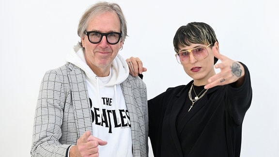 Die Podcast-Hosts Udo Dahmen mit schwarzer großer Brille und beitem Beatles-Hoodie und Sängerin Mine mit schwarzem Shirt und goldener Brille, die die Hand in die Kamera hält.