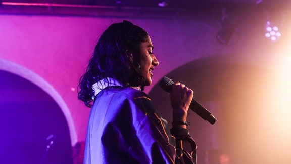 Wa22ermann bei einem Konzert: Die Rapperin hält ein Mikrofon und steht auf einer lila erleuchteten Bühne