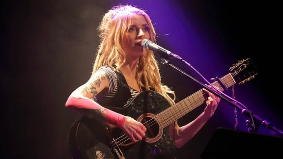 Musikerin Sarah Lesch steht mit Gitarre in der Hand am Mikro und singt. 