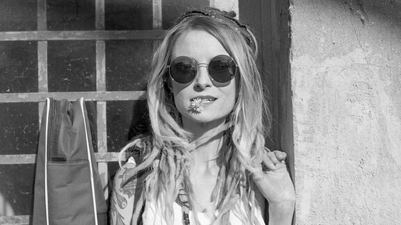 Schwarz-weiß Porträt der Musikerin Sarah Lesch mit einer runden Sonnenbrille und einer Gänseblume im Mund. 