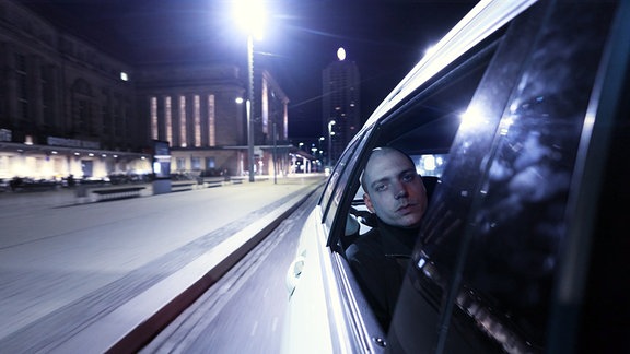 Ein Mann sitzt an einem offenen Autofenster und fährt durch die nächtliche Stadt.