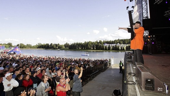 Rechts im Bild eine Konzertbühne mit einem Musiker, davor jubelndes Publikum. Im Hintergrund ein See.