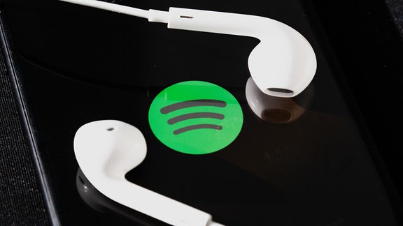 Kopfhörer liegen auf dem Bildschirm eines Smartphones, auf dem das Logo vom Musik-Streaming-Dienst Spotify angezeigt wird.