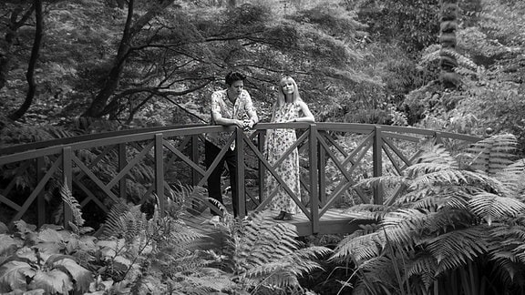Schwar-weiß-Bild eines Mannes und einer Frau, die im Wald auf einer kleinen Brücke stehen. 