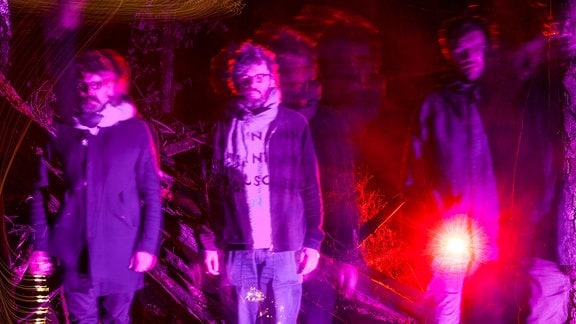 Die drei Bandmitglieder von The Notwist stehen draußen nebeneinander in pinkfarbenem Licht. Das Bild zeigt Langzeitbelichtungseffekte.