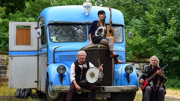 Die Musiker Kai Sichtermann und Funky K. Götzner und Musikerin Birte Volta posieren mit ihren Instrumenten vor einem alten blauen Wohnmobil. 