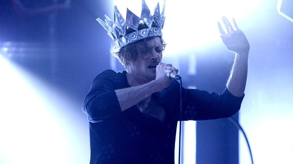 Ein Mann mit einer übergroßen Krone singt in ein Mikro.
