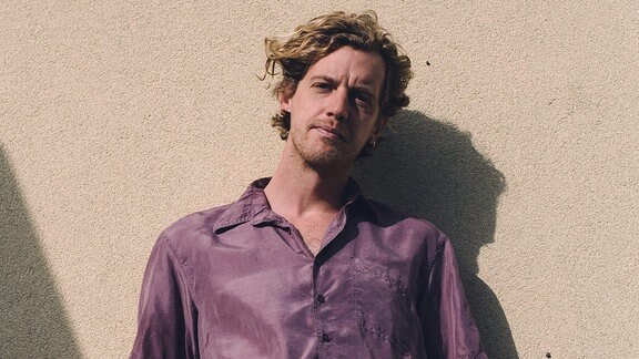 Musiker Maarten Devoldere alias Warhaus steht in der Sonne und lehnt an einer Hauswand.