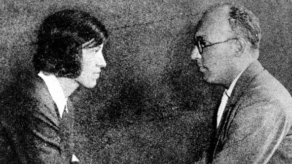 Eine Frau mit dunklen Haaren und ein Mann mit Brille sitzen sich gegenüber und sehen sich an.