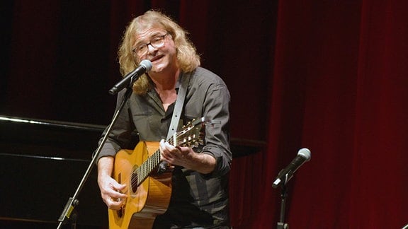 Ein Mann mit halblangen Haaren und Brille steht auf einer Bühne vor einem Mikrophon und spielt Gitarre.