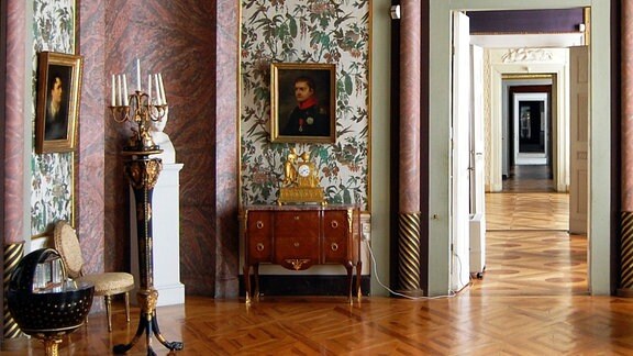 Blick in ein Prunkzimmer mit Schmuck-Tapeten, gemalten Porträts, Kronleuchter. An der Decke sind Reliefs in Form von Sonne und Sterne.