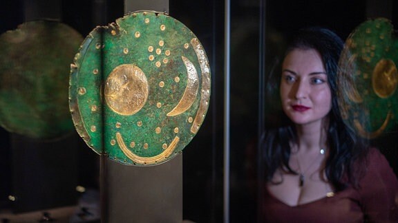 Eine Frau schaut in einen Glaskasten mit einer einer grünen Metallscheibe mit goldenen Elementen.