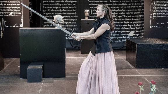 Eine Frau mit schwarzer Weste und rosa Rock streckt ein Schwert nach vorn. Im Hintergrund ist Text mit weißer Schrift auf schwarze Stelen geschrieben und Büsten stehen auf schwarzen Sockeln.
