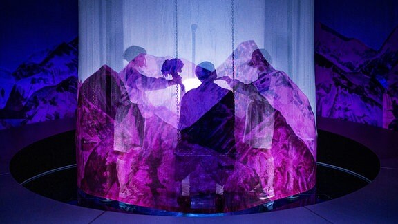 Auf einer in lila Licht getauchten Bühne stehen verhüllte Figuren in einem Kreis und werfen Schatten. 