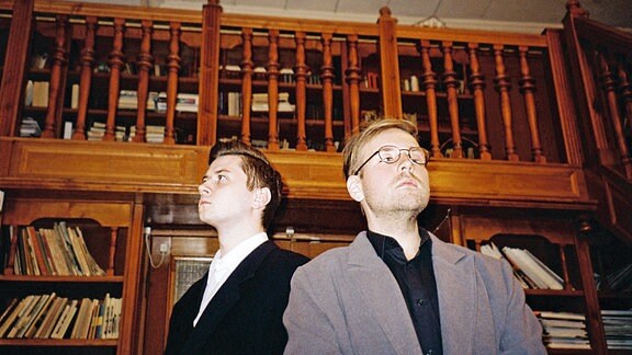 Zwei junge Männer stehen mit aufgesetztem Blick in einer Bibliothek