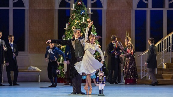 Eine Frau in weißem Kleidchen lässt sich von einem Mann auf der Bühne mit Weihnachtsbaum beim Tanz drehen.