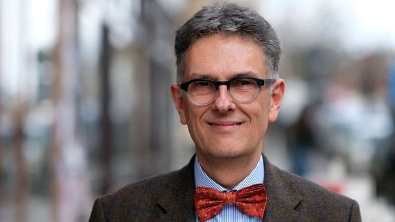 Oliver Zille, Direktor der Leipziger Buchmesse, ein Mann mit grauen haaren und Brille, er trägt einen Anzug und eine rote Fliege.
