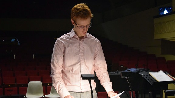 Ein Mann in einen rosanen Hemd steht am Dirigentenpult und blättert durch die Partitur.