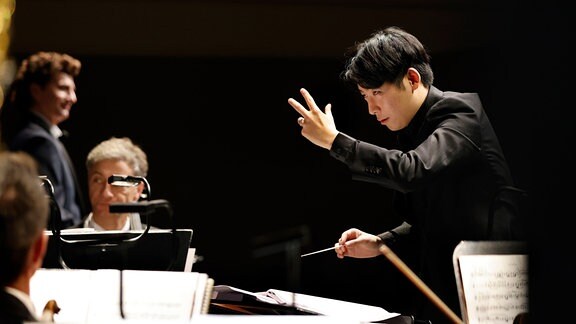 Ein Mann im Anzug steht vor einem Orchester und gestikuliert mit der linken Hand.