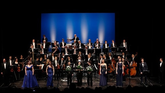 Vor einem Orchester in Anzügen stehen mehrere Personen in blauen Kleidern und Anzügen auf einer Bühne