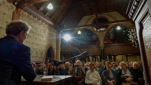 Prof. Dirk Oschmann hält eine Lesung vor Publikum in einer Kirche.