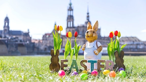 Ein Osterhase mit einem Schild Easter, Tulpen und bunten Ostereiern am Königsufer Dresden mit Blick auf die Katholische Hofkirche.