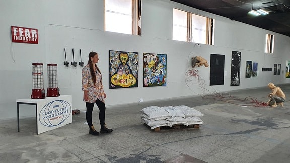 Eine Frau steht in einem Industriebau mit Bildern an den Wänden und unterschiedlichen Kunstobjekten auf dem Boden.