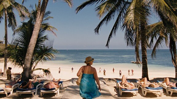 Eine Frau mit Hut und Strandtuch bekleidet steht unter Palmen und blickt aufs Meer.