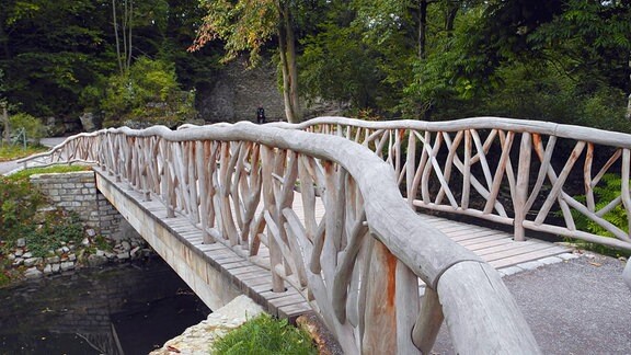 Holzbrücke mit kunstvoll geschnitztem Geländer führt über einen Fluss 