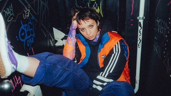 Die Musikerin Paula Carolina sitzt mit angewinkelten Bein auf einer mit Graffiti beschmierten Clubtoilette