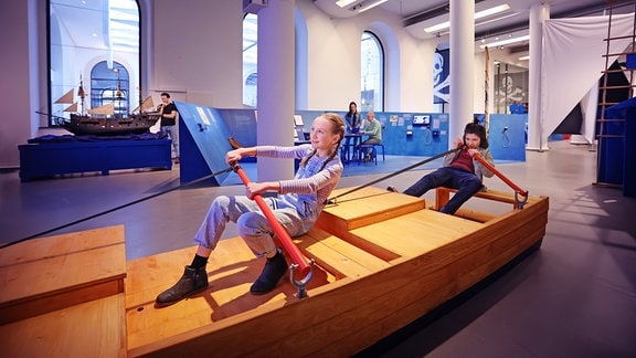 in einem Ausstellungsraum sitzen zwei Kinder in einer hölzernen Boot-Modell und Rudern.
