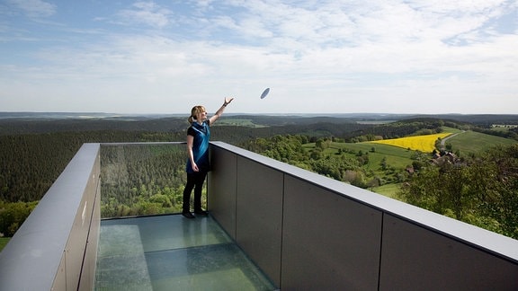 Eine Frau wirft einen Porzellanteller über die Brüstung einer Aussichtsplattform, die in ein grünes Tal hineinragt.