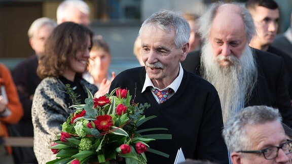 Ein Mann mit kurzen weißgrauen Haaren steht mit einem Blumenstrauß zwischen mehreren Menschen.