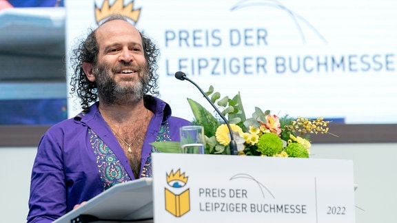 Ein Mann mit lila Hemd steht mit einem Blumenstrauß an einem Rednerpult.