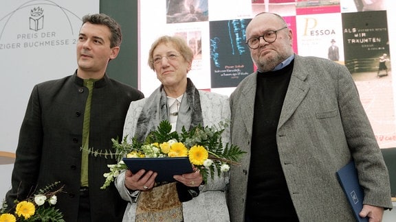 Drei Personen mit Blumen und Mappen posieren für ein Gruppenbild.