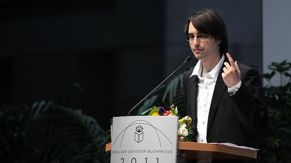 Ein Mann mit schwarzem Jackett und weißem Hemd steht an einem Rednerpult.