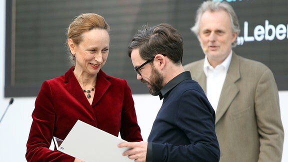 Ein Mann in schwarzem Hemd schaut in eine weiße Mappe. Dahinter stehen ein Mann im beigen Jackett und eine Frau mit rotem Blazer.