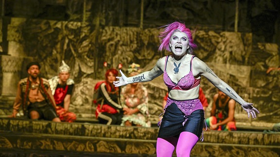 Eine Frau mit pinken Haaren und einem pinken BH steht auf einer Bühne und schreit. 