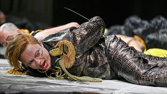 Ein Schauspieler mit rötlichen Haaren und einem schwarzen Kostüm liegt auf dem Boden und schreit. 