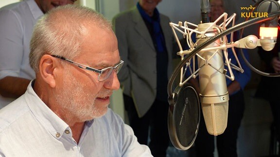 Ein Mann sitzt vor einem Mikrofon in einem Studio, er trägt ein helles Hemd und eine Brille. 
