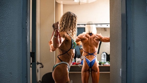 Zwei Bodybuilderinnen in Bikinis machen Dehnübungen vor einem Spiegel. 
