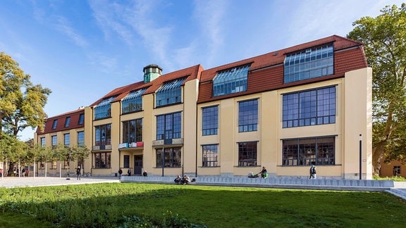 Die Bauhaus-Universität Weimar vor blauem Himmel ist zu sehen, vor dem langgestreckten Gebäude mit vielen Fenstern befindet sich eine Wiese, links und rechts vom Gebäude stehen Bäume.