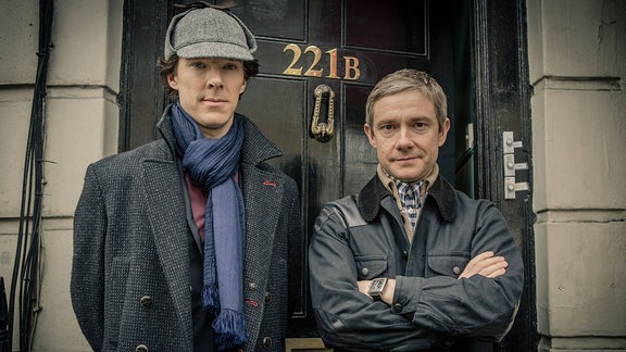 Benedict Cumberbatch und Martin Freeman: Ein Mann mit Jagdhut und ein anderer Mann mit Wetterjacke stehen nebeneinander vor einer Tür.