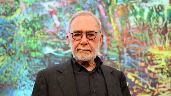 Ein als der Maler Gerhard Richter bekannter älterer Mann mit Brille, grauem Haar, grauem Bart und einem dunklen Anzug steht vor einer bunten Wand 