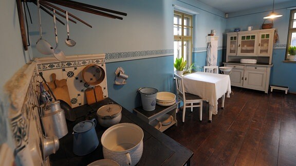 Eine in Blautönen gestrichene Küche mit einem alten weißen Schrank, kleinem weißen Tisch mit zwei Stühlen und im Vordergrund alten Küchen-Utensilien auf einem Herd