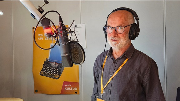 Ein älterer Mann steht mit Kopfhörern vor einem Radiomikrofon. 