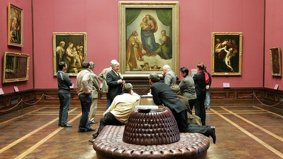 Eine Ausstellungsraum voller Besucher mit der Sixtinischen Madonna und anderen Gemälden, mitten im Raum eine Sitzmöglichkeit